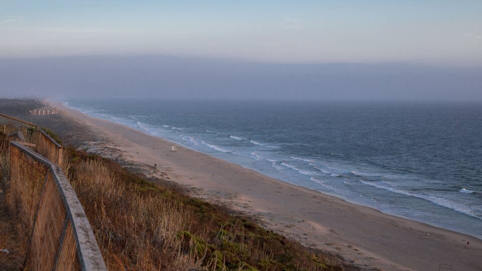 A coastal landscape with a sandy beach, ocean waves, and a wooden fence.