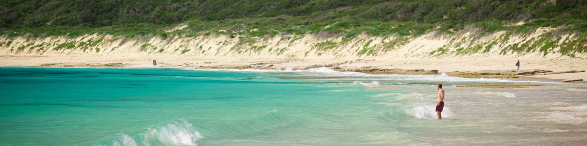 A beach with clear turquoise water, a person standing in the water, and a green hillside in the background.