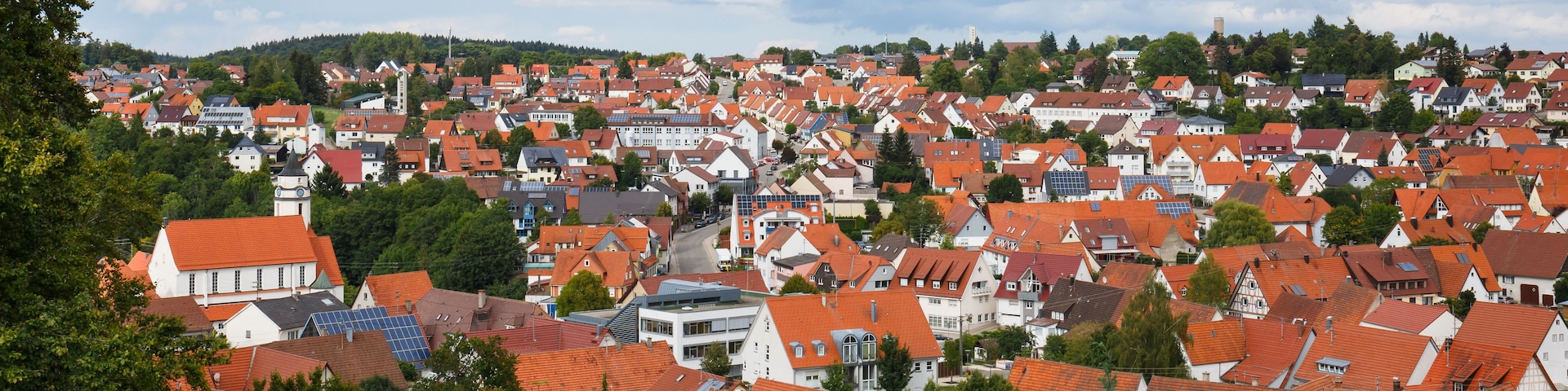 A densely packed town with red-tiled roofs, surrounded by greenery and under a partly cloudy sky.