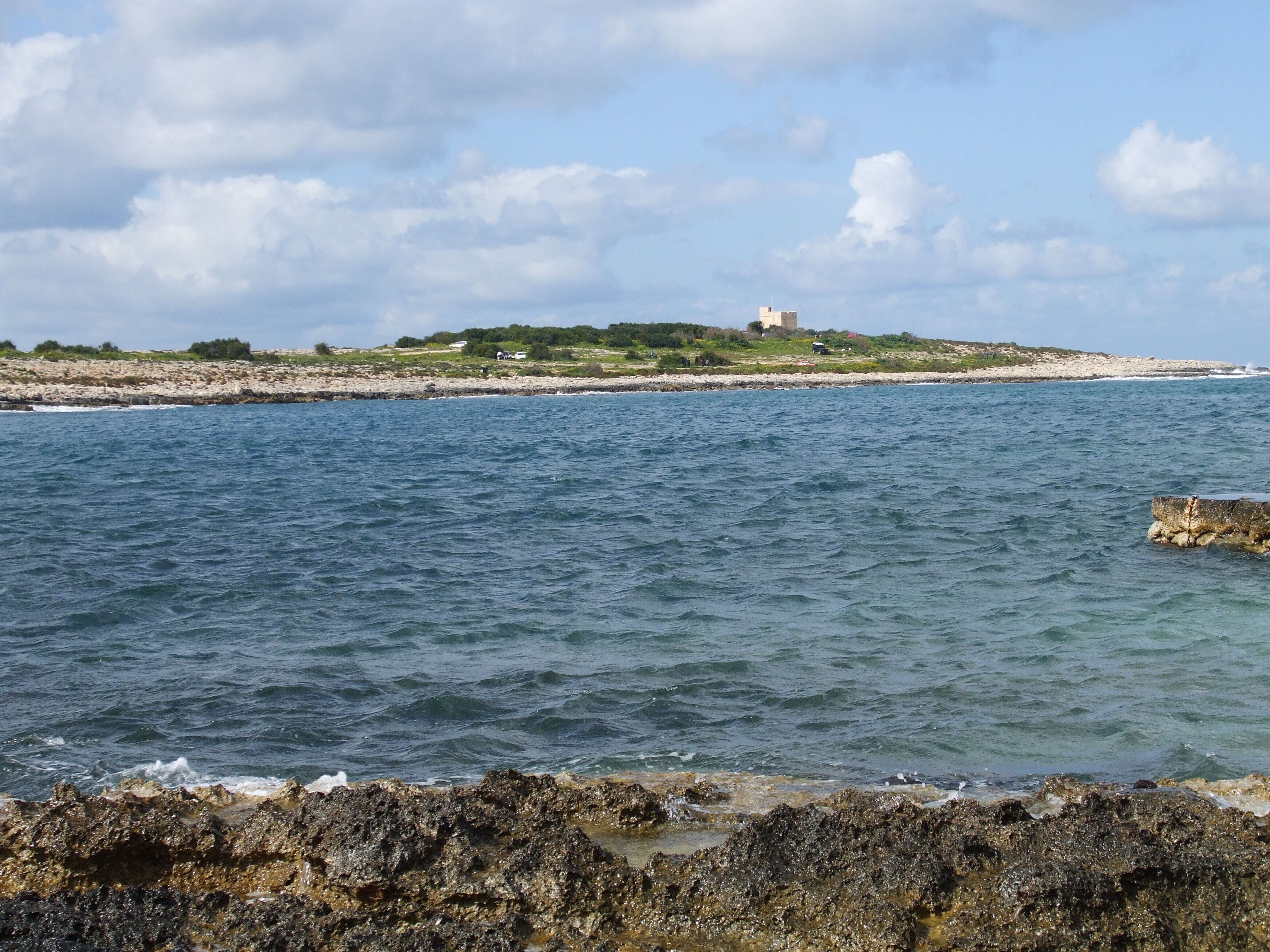 A coastal landscape with rocky shores, clear blue water, and a distant hill with a building.