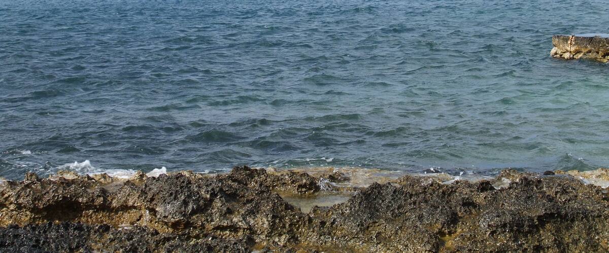 A coastal landscape with rocky shores, clear blue water, and a distant hill with a building.