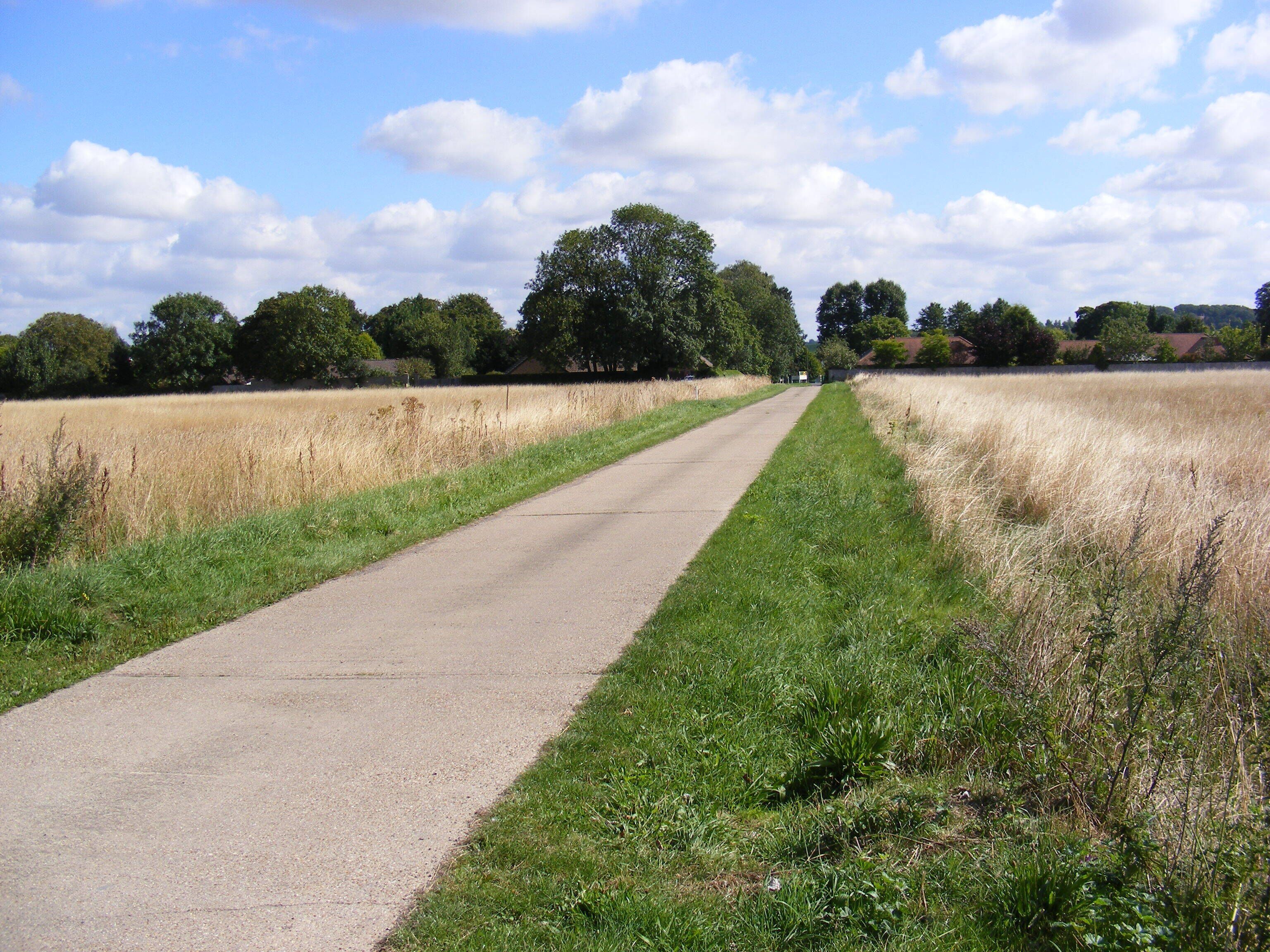 A rural road flanked by fields and trees under a partly cloudy sky.