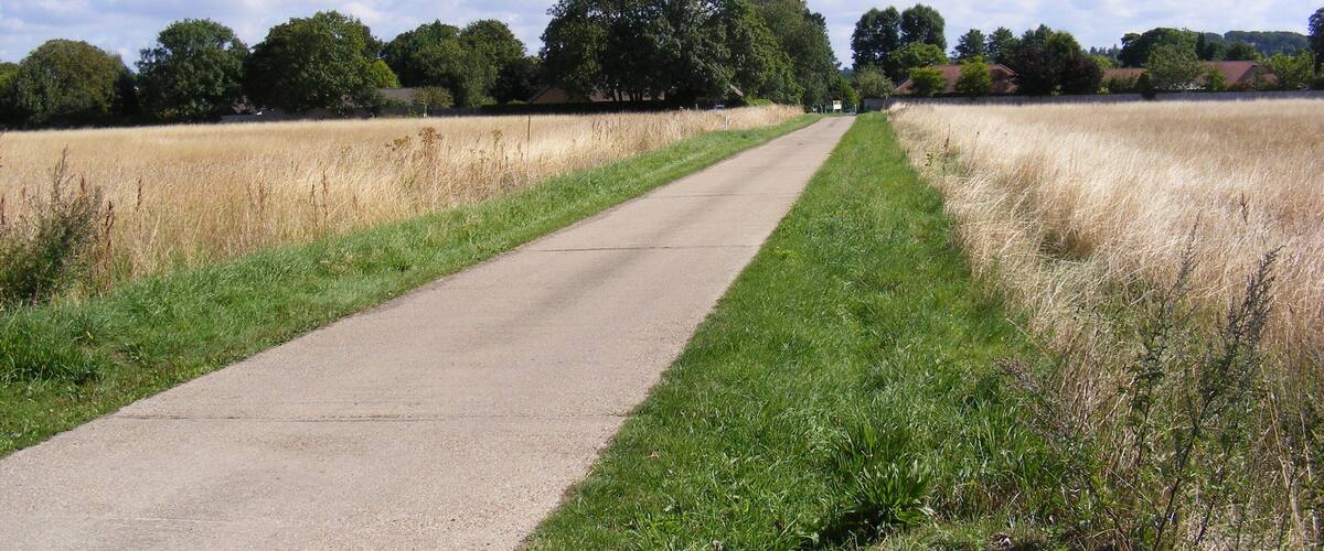 A rural road flanked by fields and trees under a partly cloudy sky.