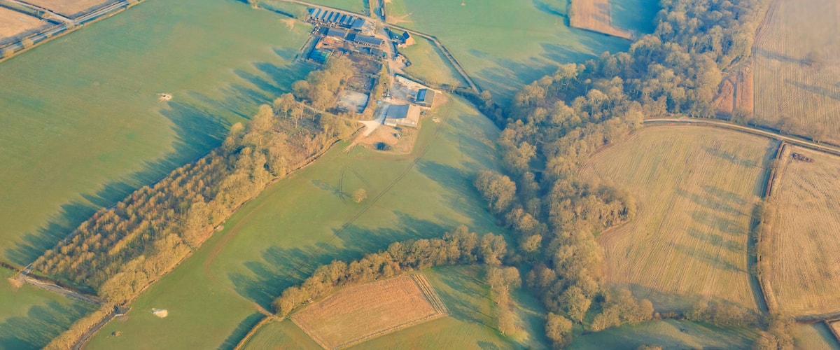 An aerial view of a rural landscape with fields, a small cluster of buildings, and a dense forest area.