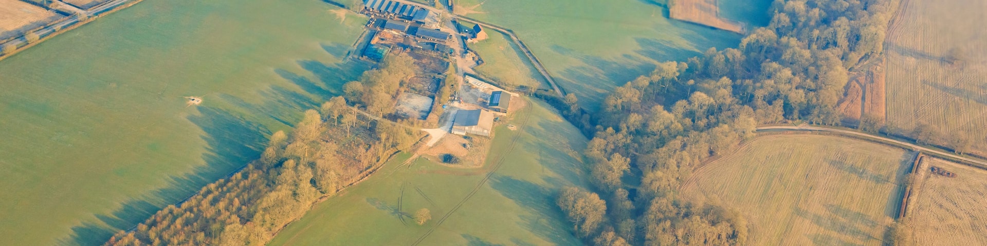 An aerial view of a rural landscape with fields, a small cluster of buildings, and a dense forest area.