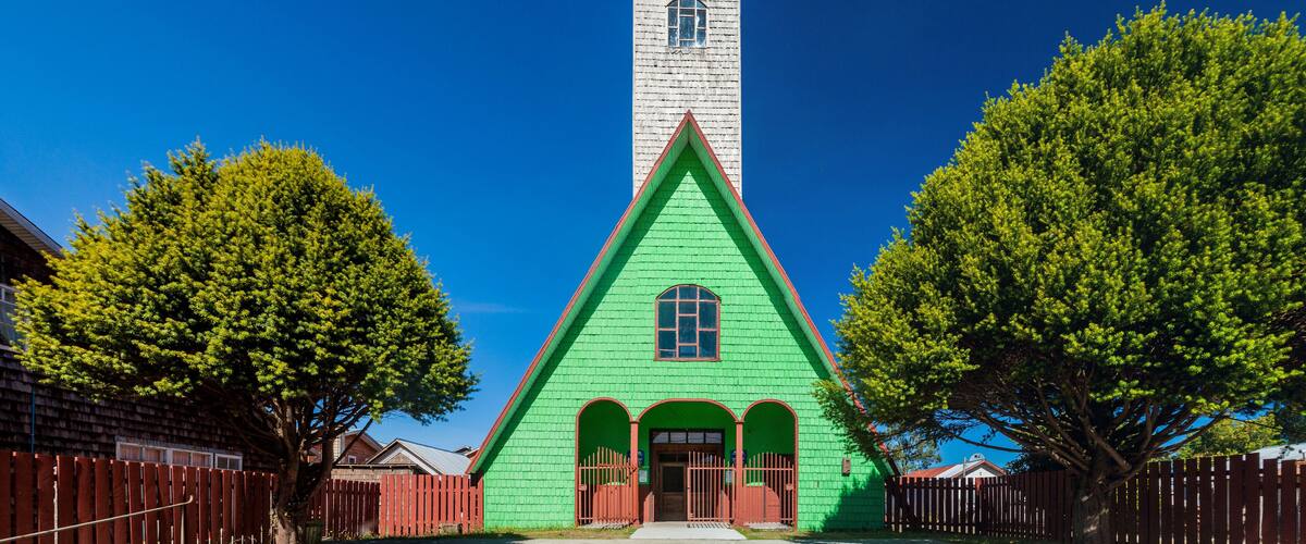 A green church with a white steeple and a cross, surrounded by trees and a red fence.