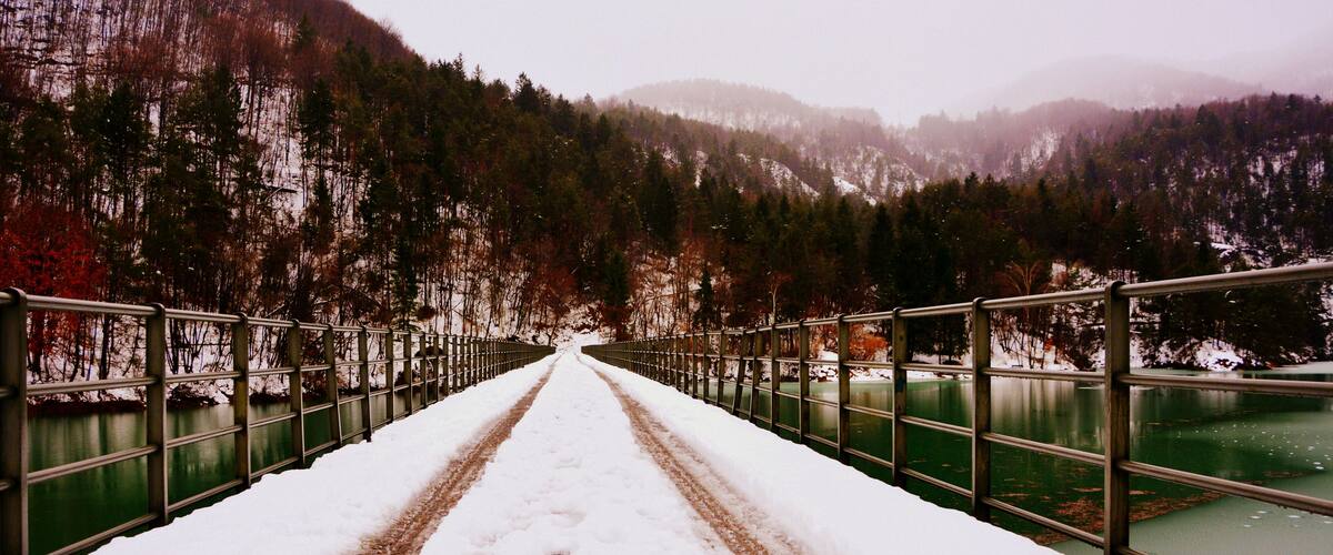 A snow-covered bridge with metal railings, leading to a forested mountain range.