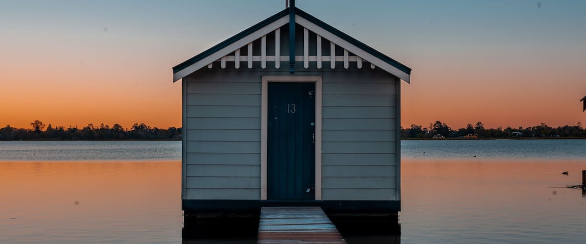 A small wooden boathouse with a dark door and a white picket fence, situated on stilts over calm water, with a sunset in the background.