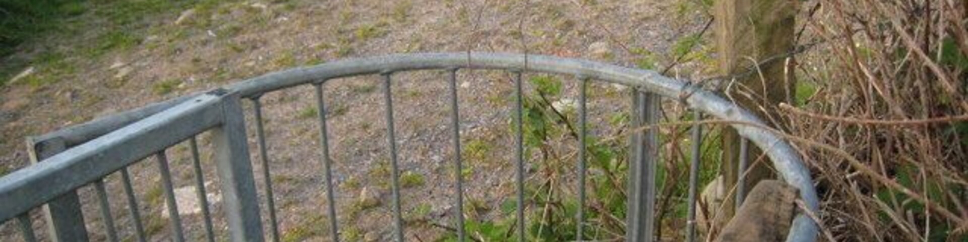 Una verja metálica en un entorno rural, con un campo cubierto de césped y una estructura de piedra al fondo.