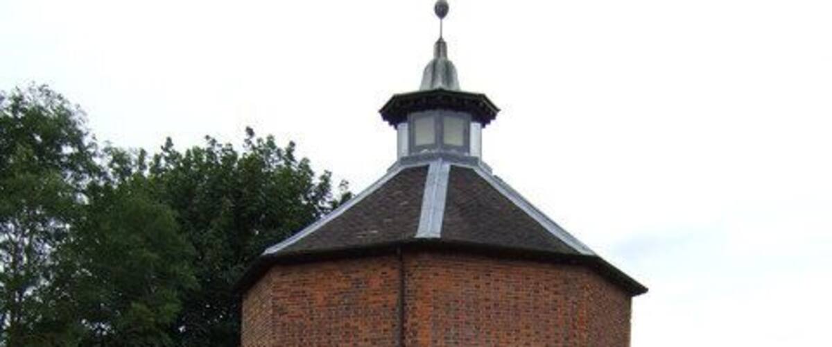 A brick tower with a conical roof and a clock, surrounded by a pond and greenery.