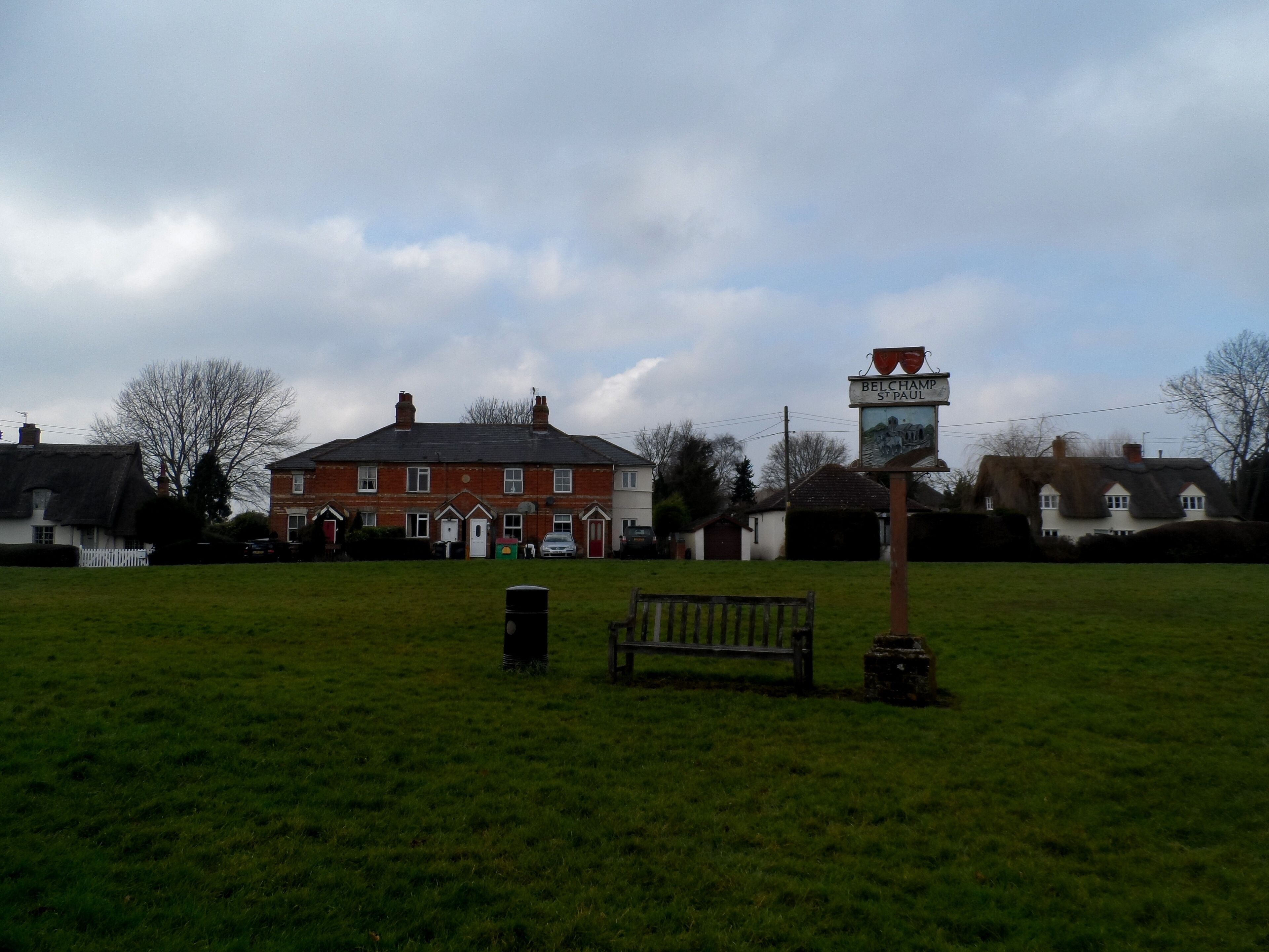 A grassy field with a bench, a signpost, and a red brick building in the background.