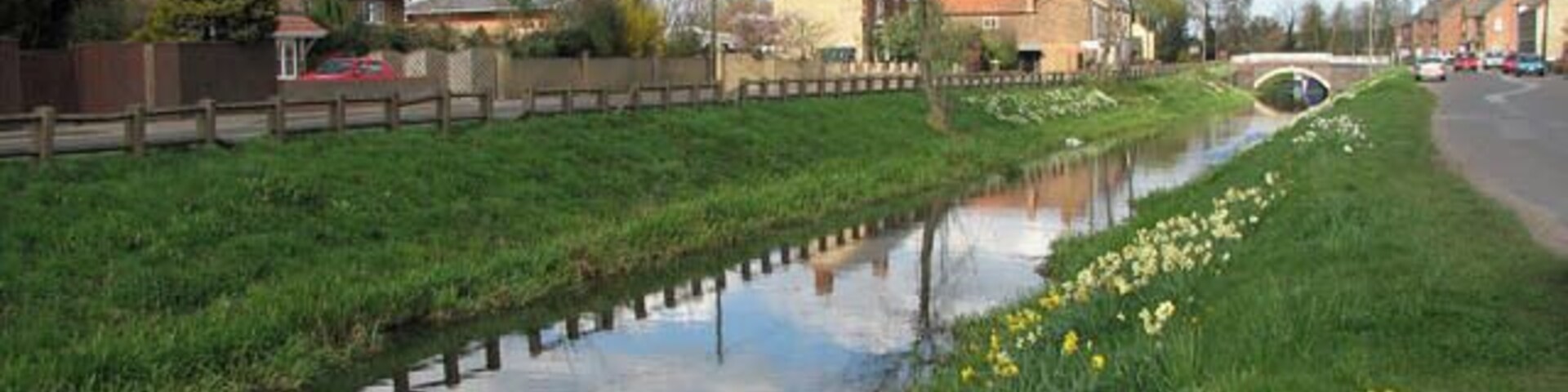 Wisbech & Upwell tramway - Goodman's Crossing Wisbech & Upwell tramway's Upwell depot > 1241222 was 5 miles 72 chains distant from Wisbech. The line had eleven sidings, with two originally allocated for passenger traffic. During the fruit season the sidings could hold more than a hundred vans. The depot had a cattle dock, a depot office, a passenger waiting room, and a coal merchant's office and staithes. There was also an ash pit, and an old GER tender was used for a water tower until 1953 when it was removed. http://www.lner.info/co/GER/wisbech/route.shtml From the Upwell depot, which was the southern terminus of the tramway, the trains headed northwards > https://www.geograph.org.uk/photo/1241251 to the village of Outwell, crossing Small Lode > https://www.geograph.org.uk/photo/1241268 and passing a farm before emerging on Low Side > https://www.geograph.org.uk/photo/1241442 which was the final request stop. A farm driveway now runs where the railway trackbed used to be > https://www.geograph.org.uk/photo/1241431. Running parallel with Well Creek (the old course of the River Nene) the tramway then ran along the grassy verge seen in the foreground before reaching the final section of reserved track at Goodman's Crossing request stop before reaching Outwell Village depot. Outwell Village depot > https://www.geograph.org.uk/photo/1241458 was located just past the bridge seen in the background - by the old course of the River Nene and adjoined by St Clement's church on the other side. It originally had four sidings and was equipped with coal chutes to transfer coal to barges for distribution through the Fens. The depot had a small office building built from red brick > https://www.geograph.org.uk/photo/1241479 and an old van body for storage. The brick office still stands, but most of the depot site has since been built over by modern housing. The name of the cul-de-sac/access road is called "The Tramway". When during the 13th century the estuary silted up, the fens surrounding the market town and port of Wisbech were reclaimed for agricultural use. The new agricultural prosperity soon attracted railways such as the Eastern Counties Railway which opened a branch in 1847. In 1848 the East Anglian Railway built a branch from Watlington. A third line was built by the Peterborough, Wisbech & Sutton Bridge Railway (later the Midland & Great Northern Joint Railway) which opened a station on the other side of the River Nene in 1866. The trains carried fruit and vegetables to markets as far afield as London. In addition, the construction of a tramway line connecting the villages Upwell and Outwell with Wisbech commenced in 1882. Board of Trade's tram restrictions resulted in the unusual-looking locomotives which not only had speed restrictions but controls at both ends and wooden cowcatchers and skirts over the wheels. Six trams a day in each direction provided passenger services, with the full one way journey taking one hour. By October 1884 traffic had grown to 3000 passengers per week, with peaks of 2000 in a day for fetes and other special events. The tram competed with a canal that ran between Wisbech and Upwell and with the canal being in a poor financial condition the tram eventually finished it off. When motor omnibuses started to appear, however, passenger numbers declined drastically and passenger services were withdrawn in 1927, whereas freight traffic continued to flourish. With the introduction of two Drewry Shunters (BR Class 04) in 1952, Wisbech & Upwell became Britain's first all-diesel line. The line closed in March 1966. http://www.lner.info/co/GER/wisbech/history.shtml