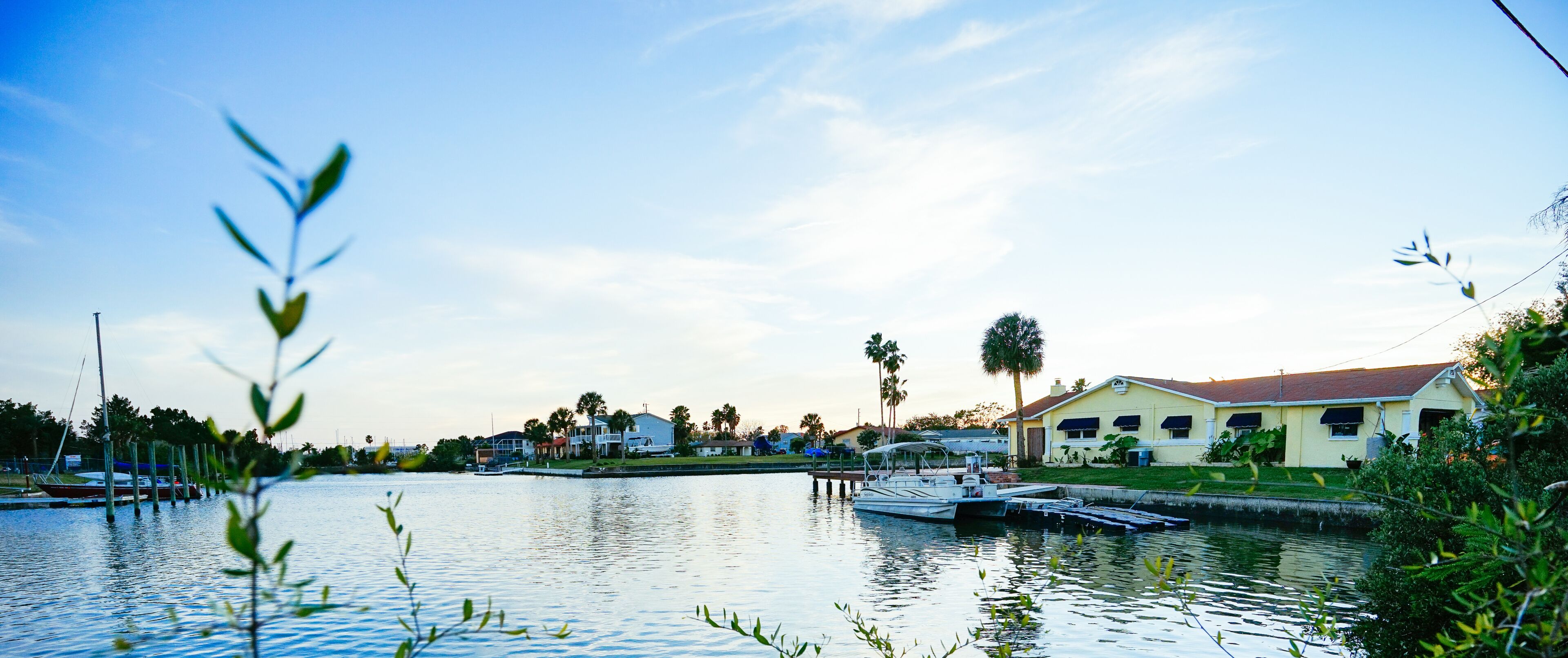 A calm waterfront with a dock, boats, a house with a red roof, and palm trees.