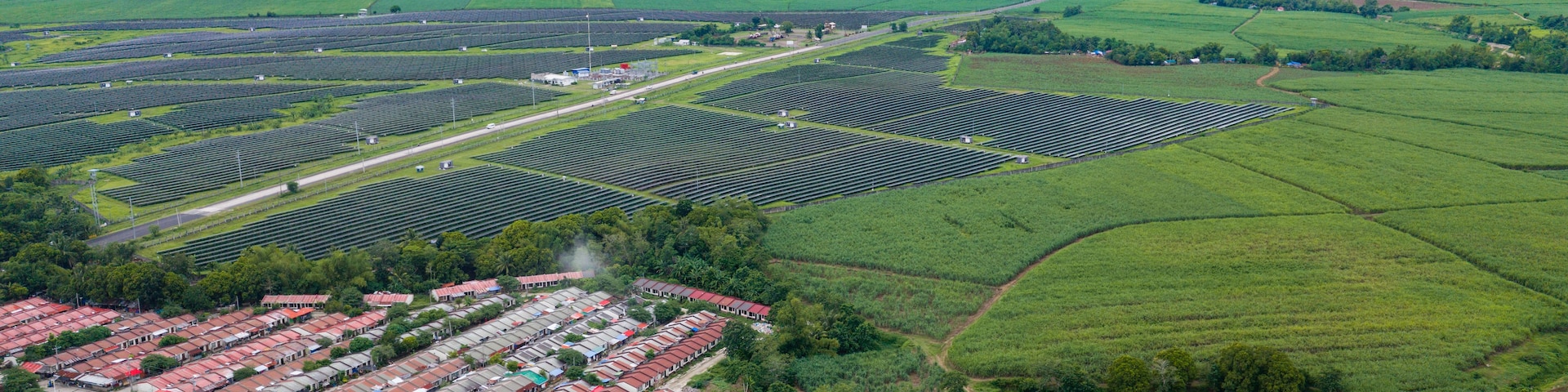 Vista aérea de una planta solar con hileras de paneles solares, una zona residencial con casas de tejados rojos y campos verdes circundantes.