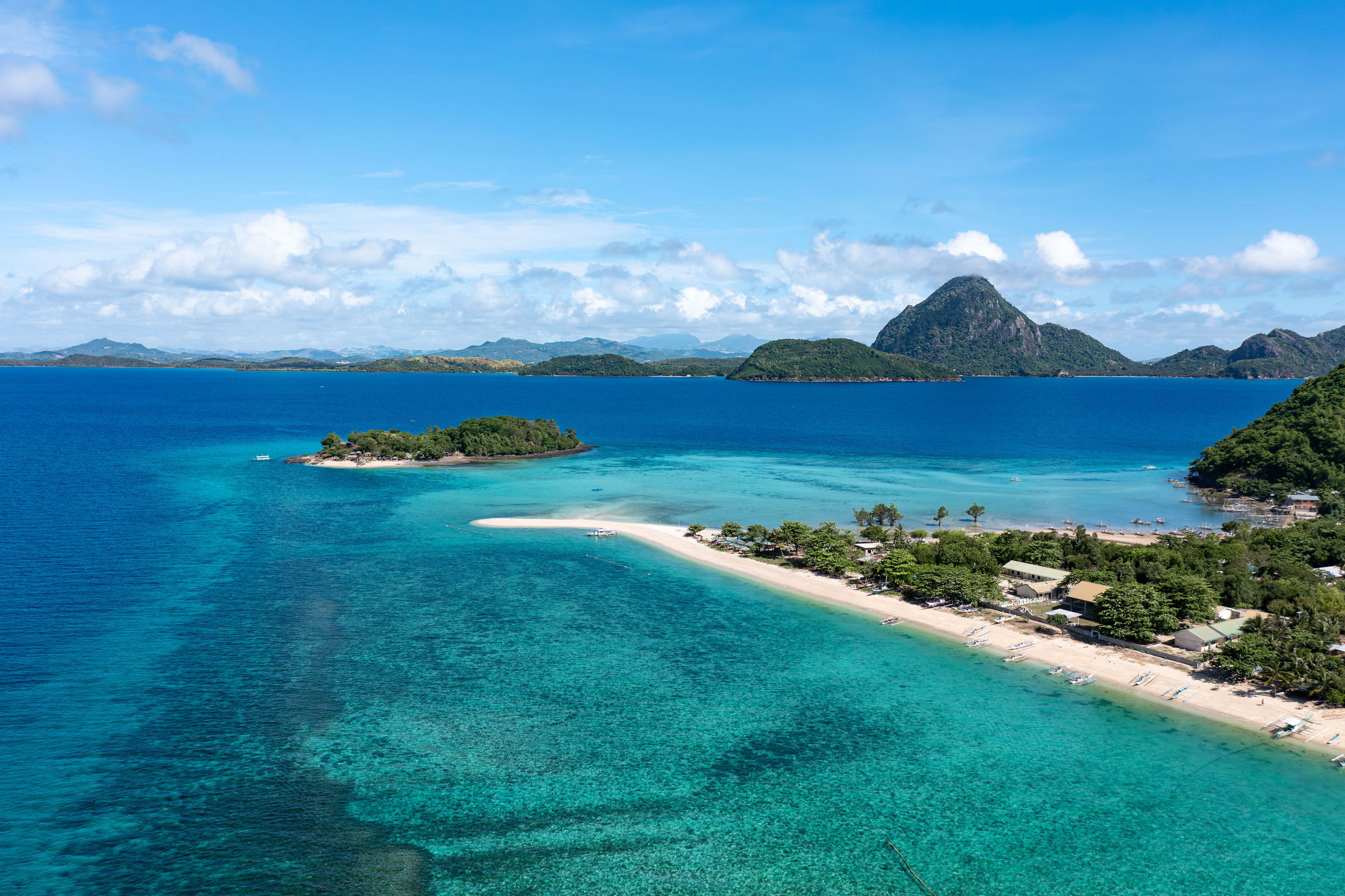 Ein tropischer Strand mit klarem türkisfarbenem Wasser, einer kleinen Insel und einer bergigen Kulisse.