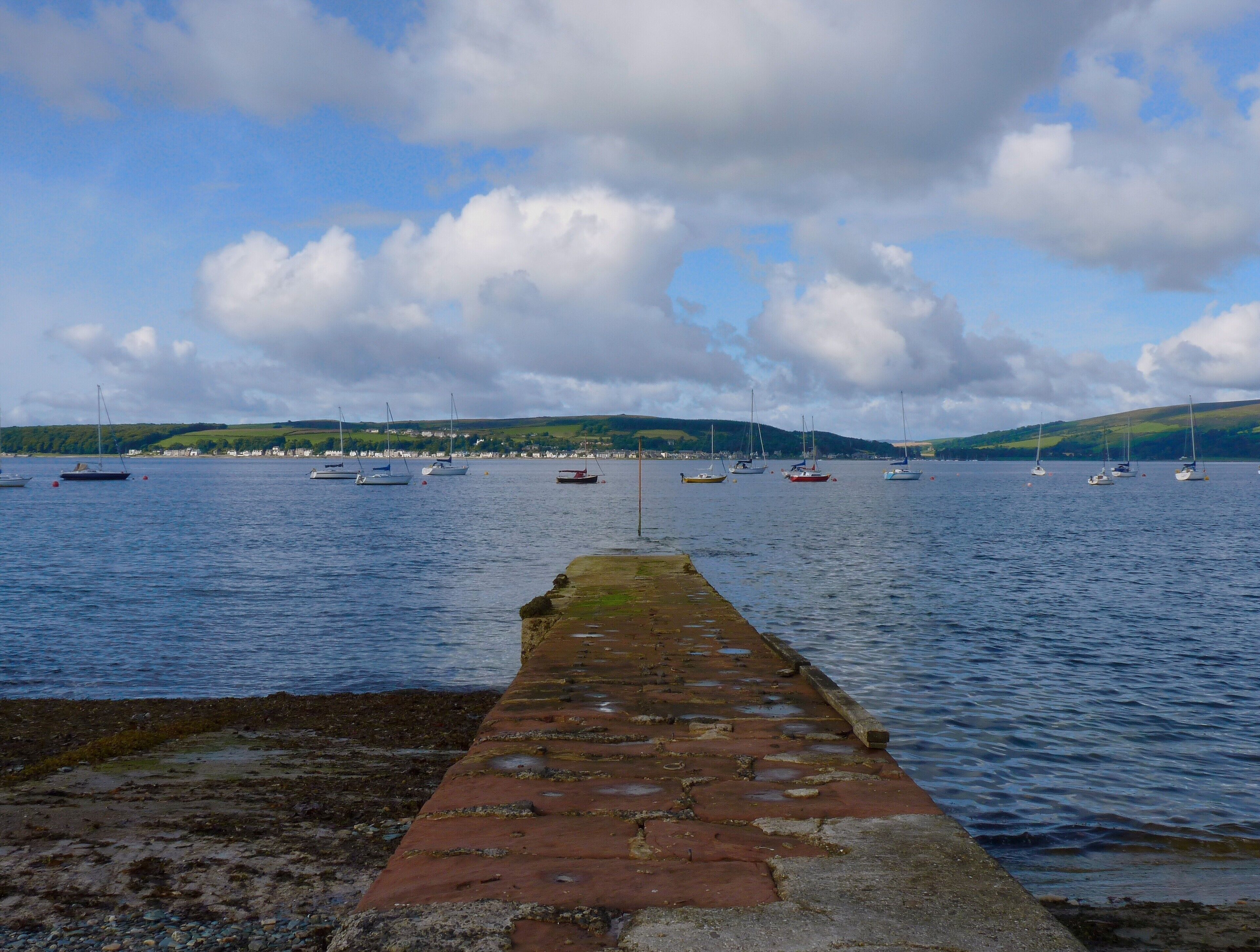 A stone pier leading into calm waters with several sailboats anchored in the distance.