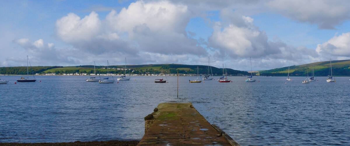 A stone pier leading into calm waters with several sailboats anchored in the distance.