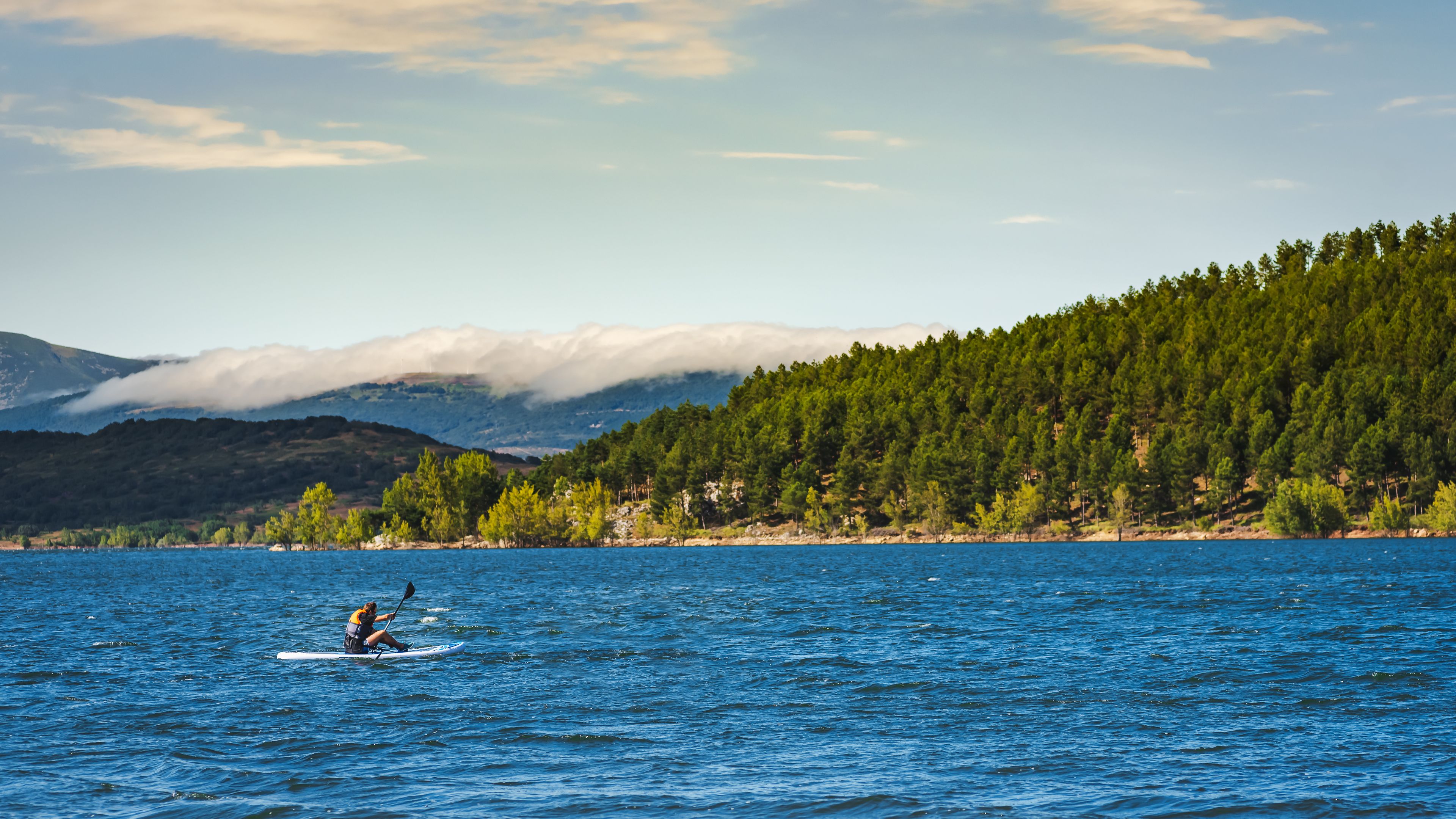 Un remer de kayak a un llac tranquil amb una serralada boscosa al fons.