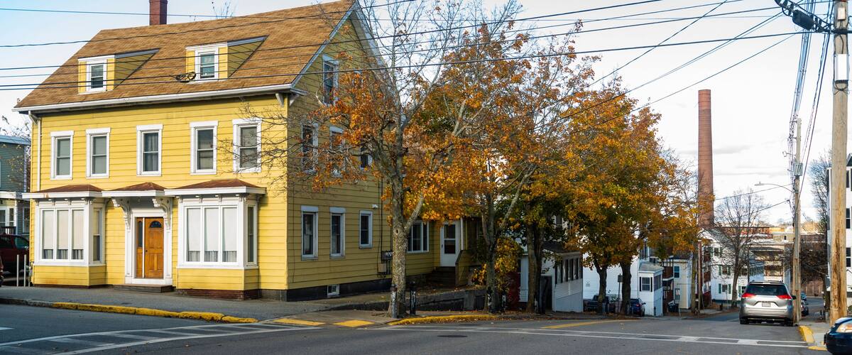 A yellow house with a brown roof, a tree with orange leaves, and a car on the street.