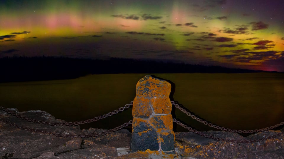 A chain-link fence with rusted posts and a chain, overlooking a calm body of water under a colorful aurora display.
