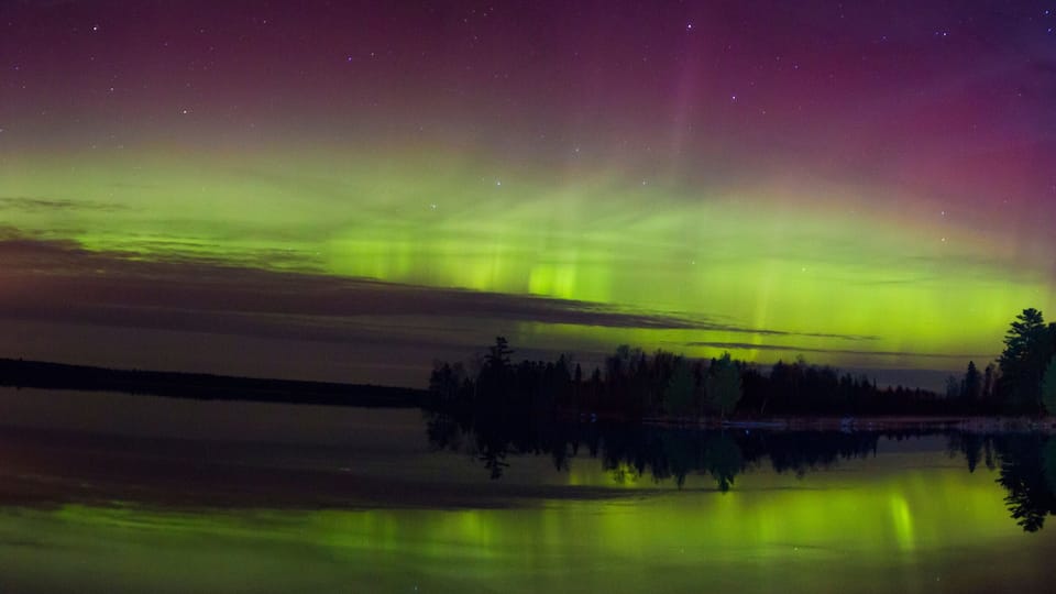 The Northern Lights over a calm lake with reflections of trees.