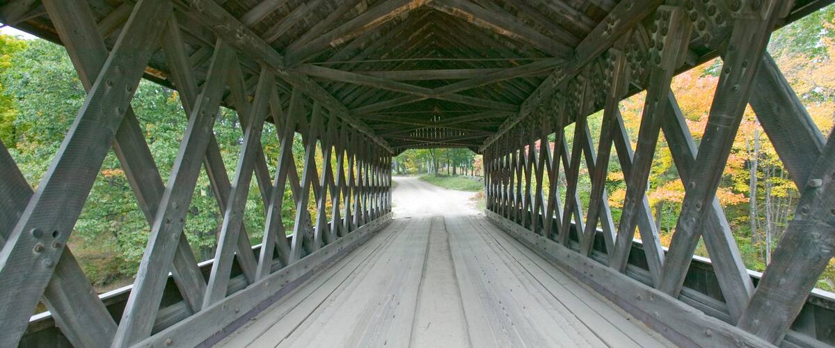 A wooden covered bridge with a metal railing, surrounded by trees with autumn foliage.