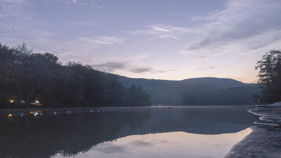 A calm lake at dusk with reflections of trees and a distant mountain range.