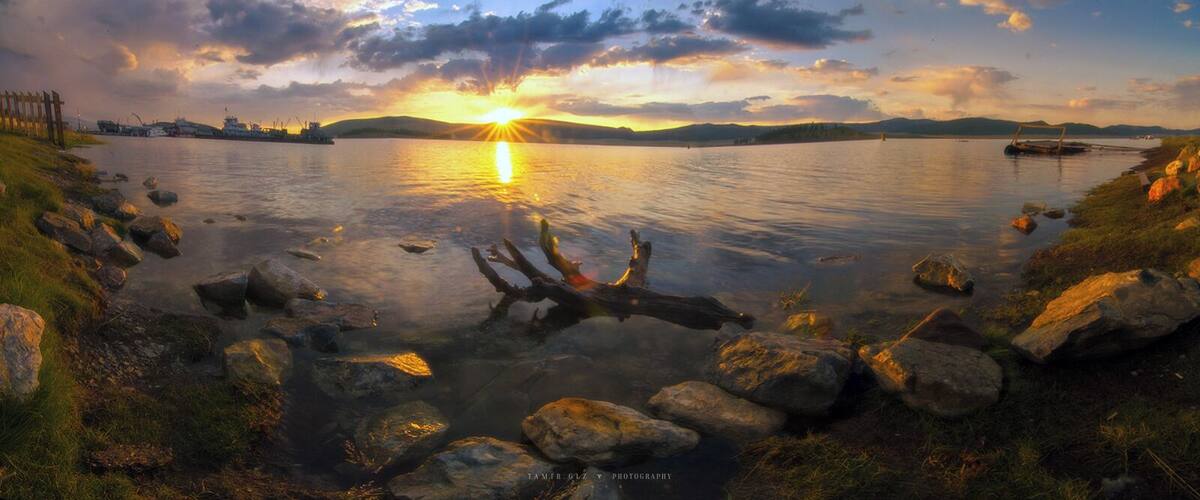 A sunset over a calm lake with rocks in the foreground and ships in the background.