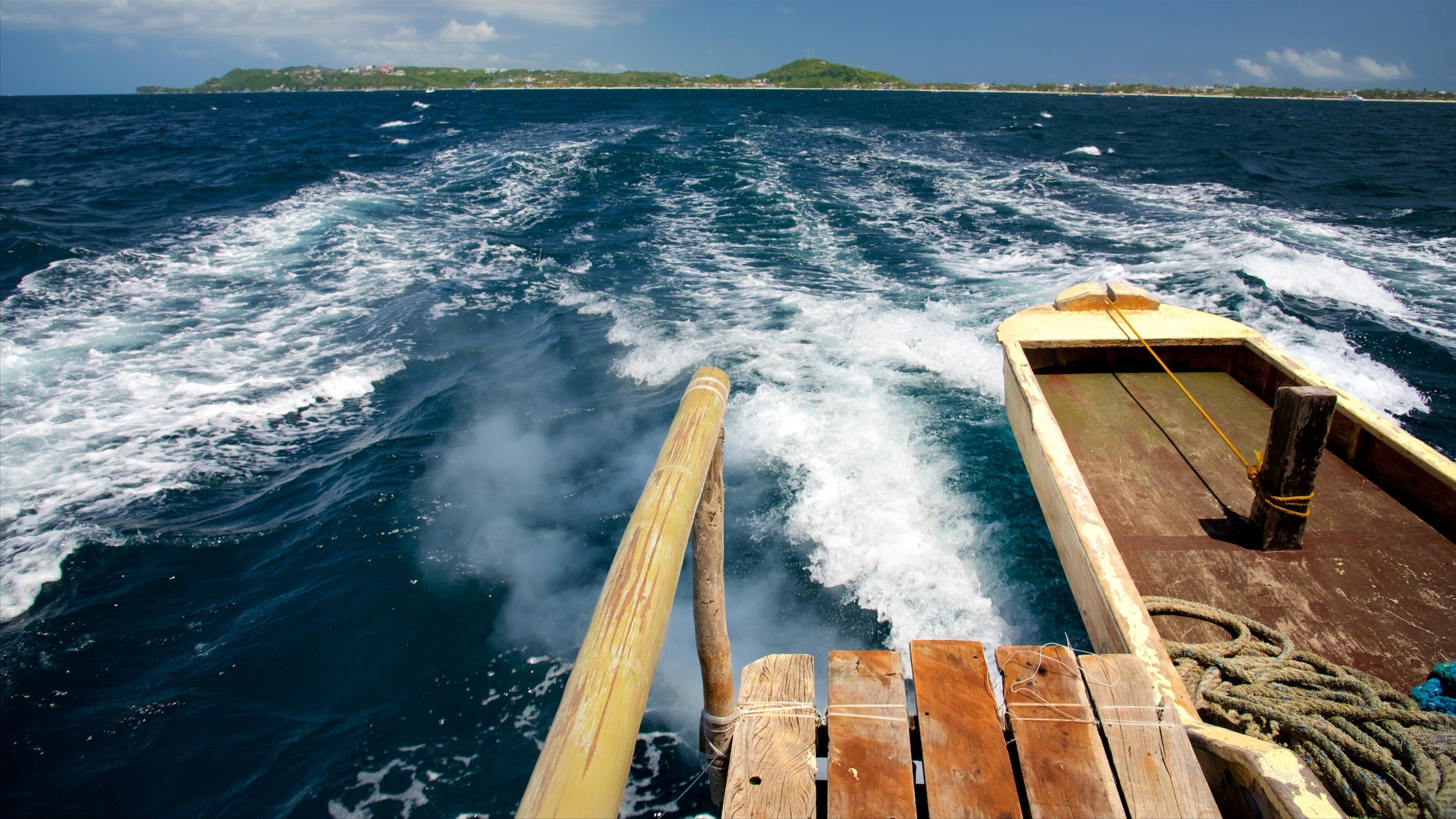 A wooden boat with a wake in the ocean, a rope, and a distant landmass with greenery and buildings.
