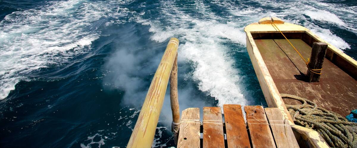 A wooden boat with a wake in the ocean, a rope, and a distant landmass with greenery and buildings.