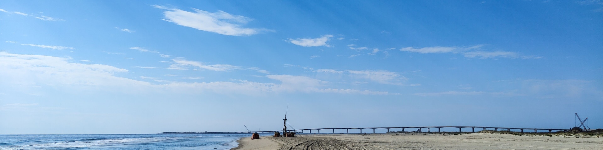 Uma praia com um píer longo que se estende para o oceano, margem arenosa e céu azul claro com nuvens esparsas.