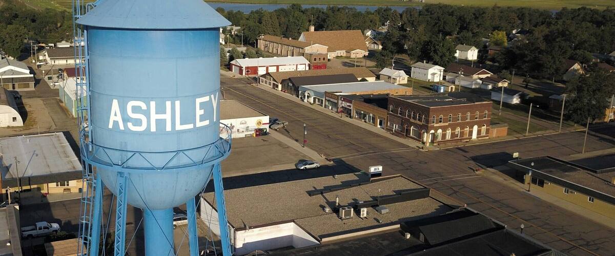 A blue water tower labeled 'ASHLEY' overlooking a small town with buildings and a river.