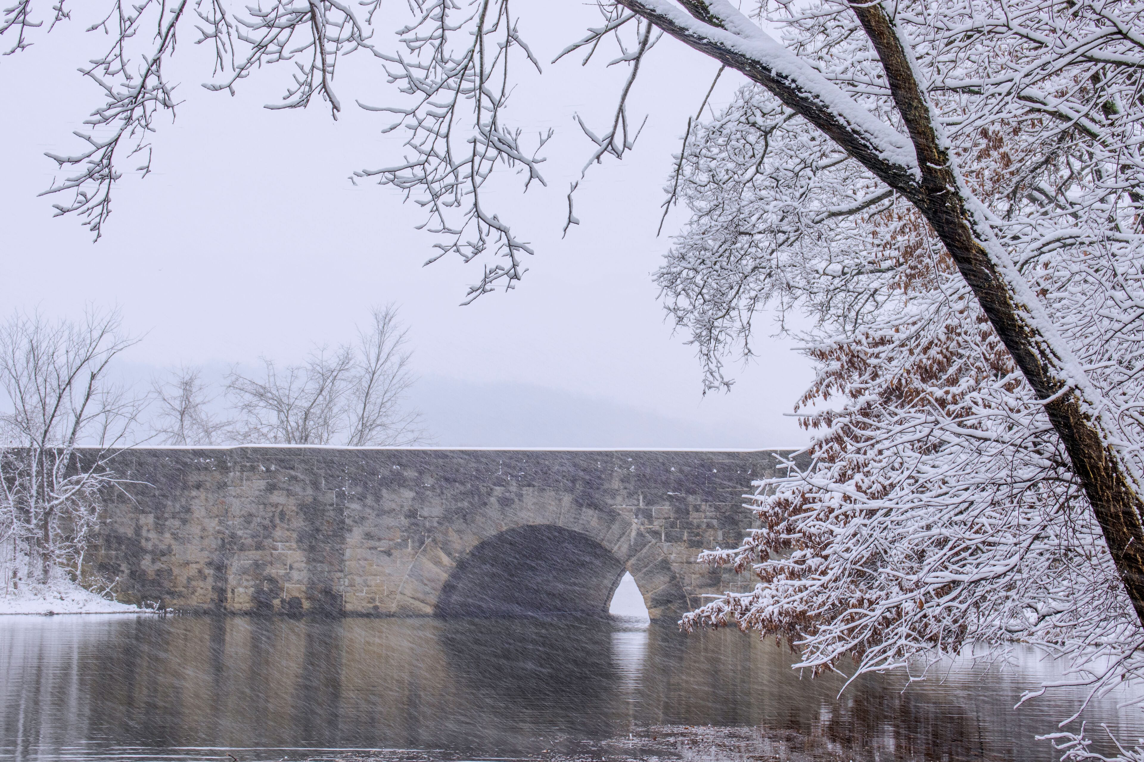 A stone bridge over a calm river, surrounded by snow-covered trees.