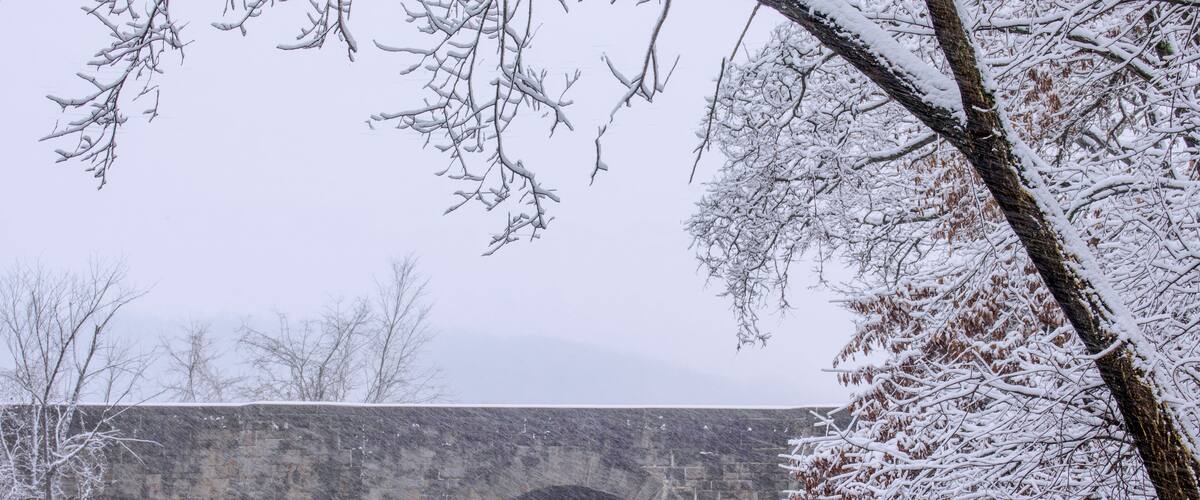 A stone bridge over a calm river, surrounded by snow-covered trees.