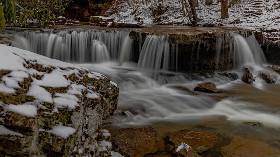 A snow-covered waterfall with rocks and trees in the background.