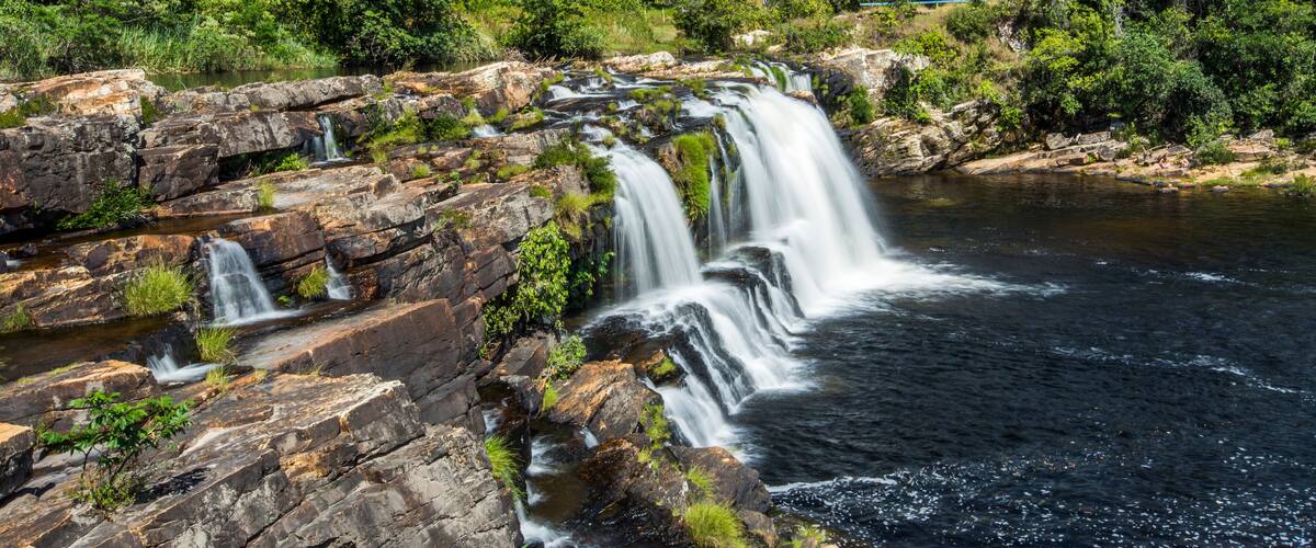 A multi-tiered waterfall cascading over rocky terrain surrounded by dense tropical vegetation.
