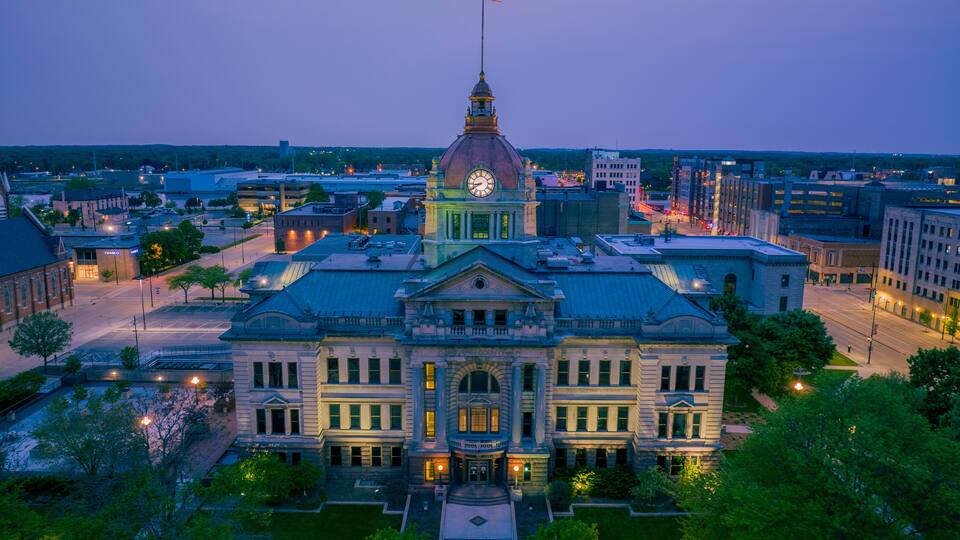 A large, historic building with a prominent dome and flag, surrounded by a well-maintained courtyard and urban streets.