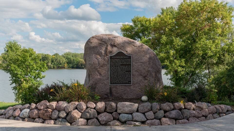 A large rock with a plaque, surrounded by a stone wall and greenery, near a lake.