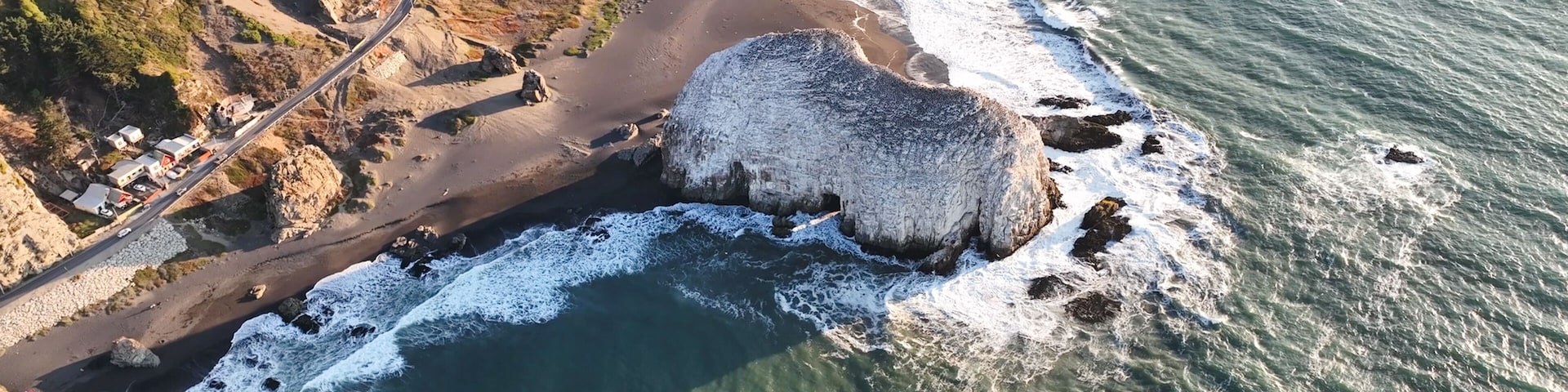 A coastal road with a rocky shoreline, a large boulder, and a building complex in the background.