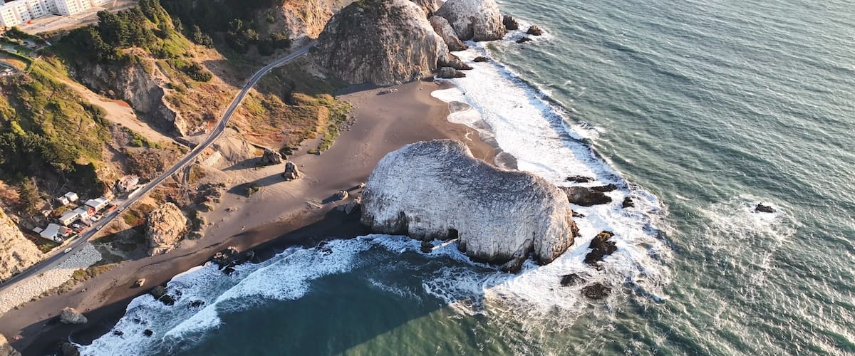 A coastal road with a rocky shoreline, a large boulder, and a building complex in the background.