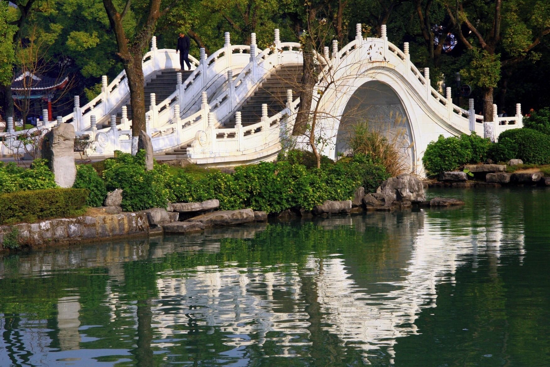 Un puente de piedra blanca tradicional sobre un estanque tranquilo, rodeado de exuberante vegetación y árboles.