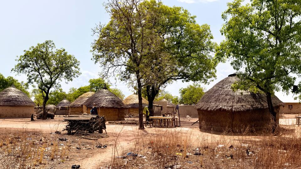 Thatched-roof huts, dry grass, and scattered trash in a rural setting.