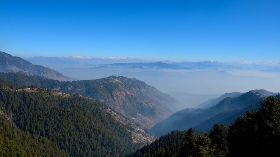 A mountainous landscape with dense forests and distant snow-capped peaks under a clear blue sky.