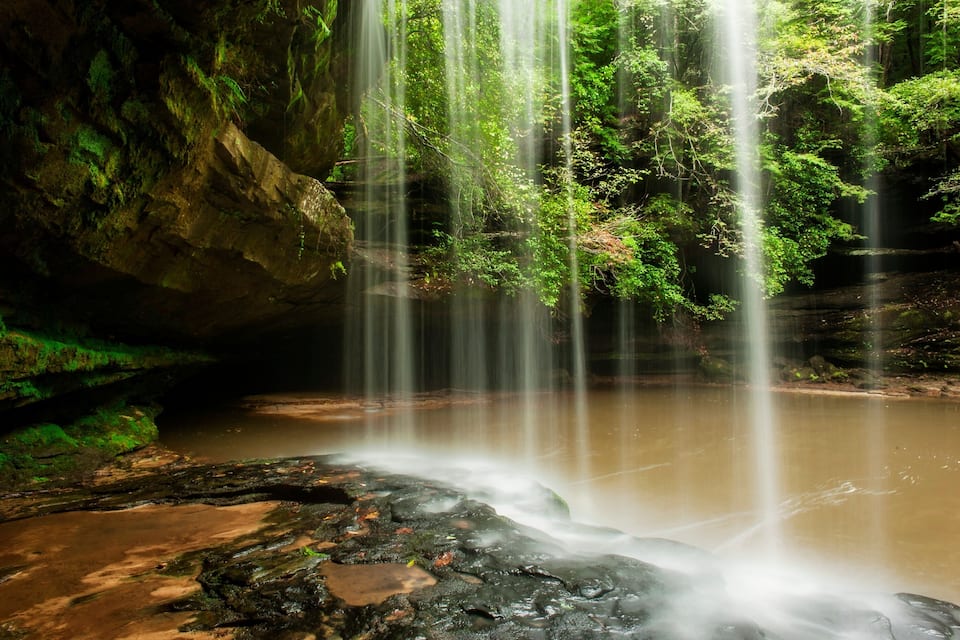 A waterfall cascading into a calm pool surrounded by lush greenery and rocky terrain.