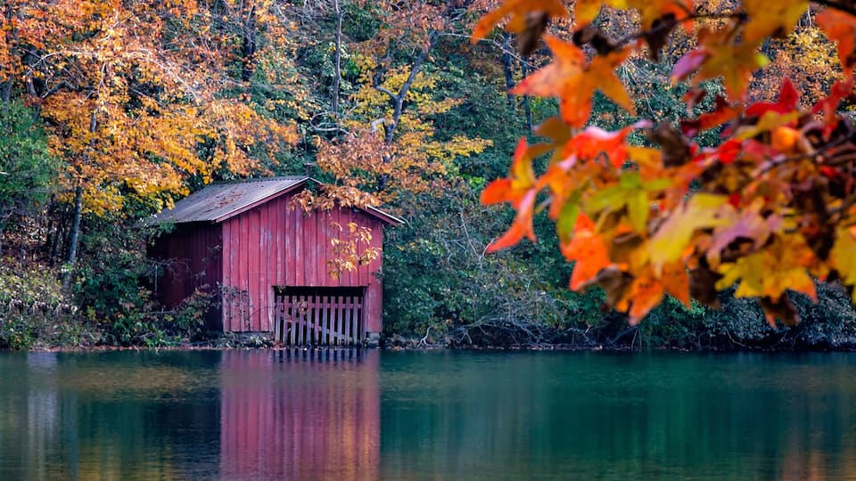 A red wooden structure by a calm lake surrounded by autumn foliage.
