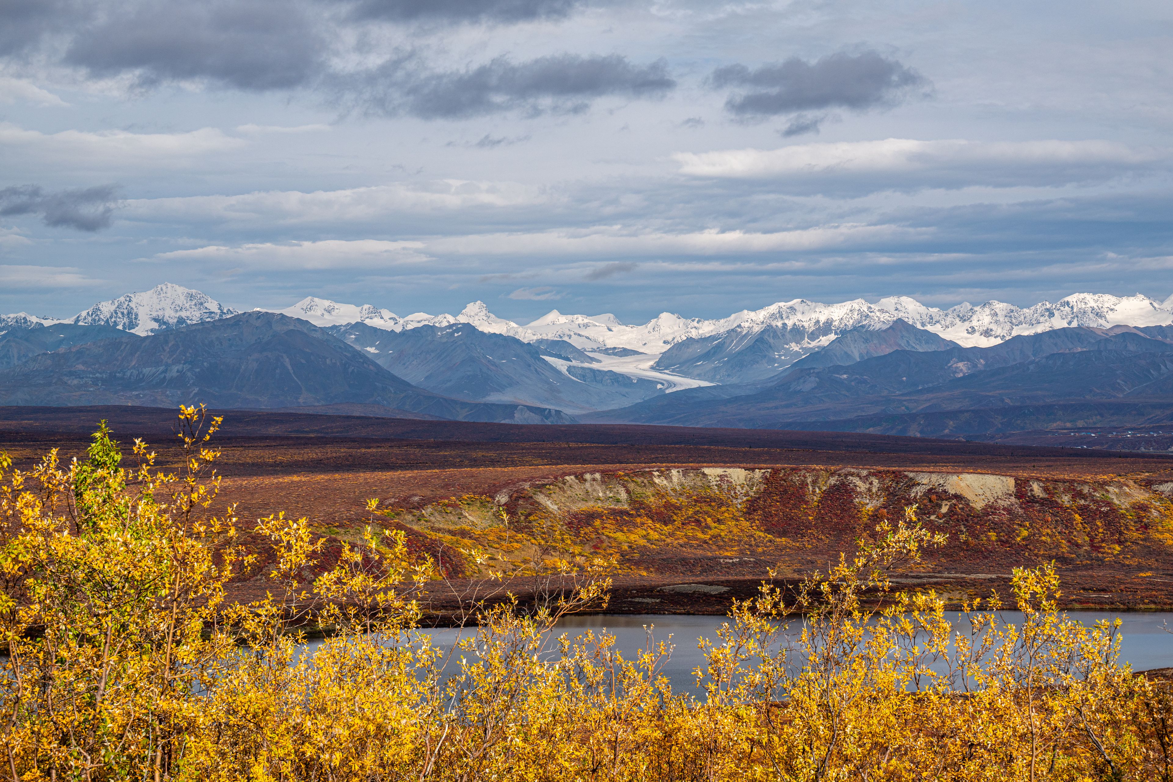 A mountainous landscape with snow-capped peaks, a river in the foreground, and autumn-colored foliage.