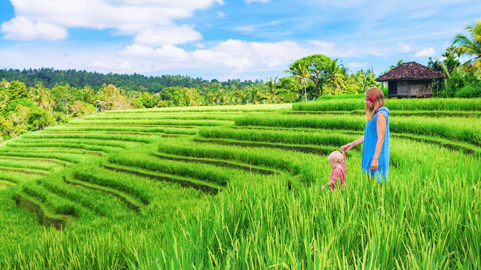 A woman and a child in a rice paddy field with terraced hills and a traditional hut in the background.