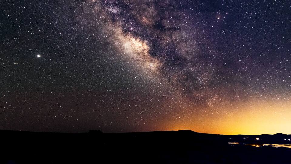 A starry night sky with the Milky Way visible, a silhouette of a mountain range, and a distant city skyline.