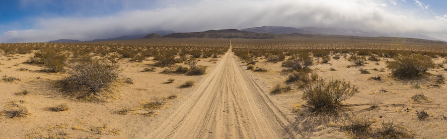 Wide panoramic view of sand road leading straight through the dry wilderness of desert.
