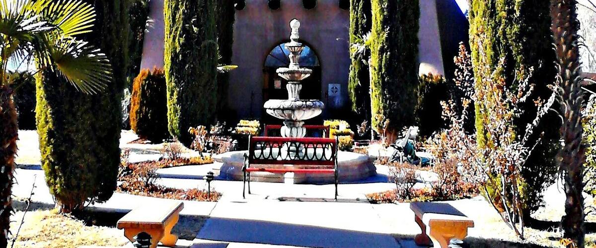 A fountain surrounded by tall cypress trees and a building with a dome and bell tower.